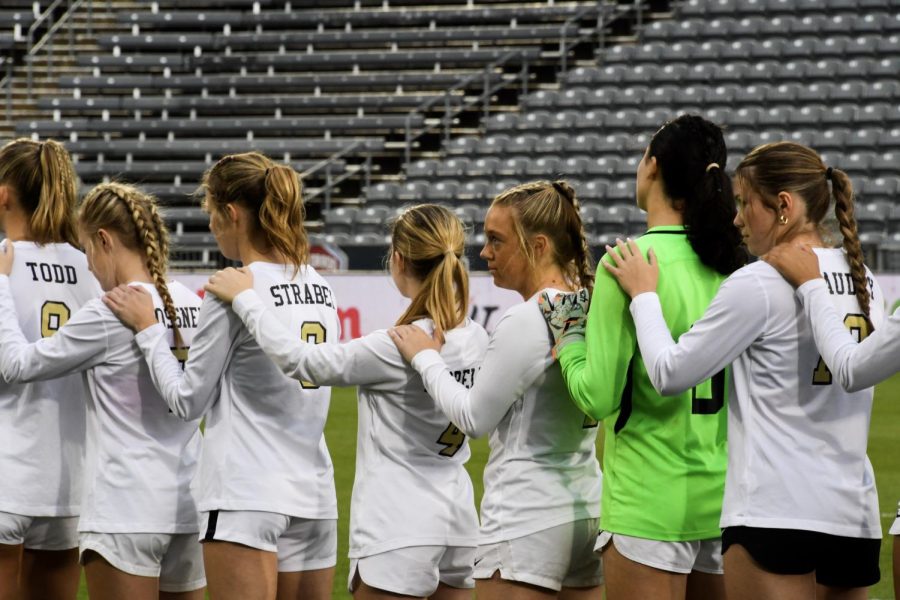 Standing in a line, the varsity girls soccer team listens to the national anthem play before the State Championship game against ThunderRidge High School (TRHS) at Dick’s Sporting Goods Park May 25. The game was delayed 30 minutes due to weather. The weather delay was definitely difficult for many of us, as it disrupted what each of our individual pregame rituals are. Visualizing a win was definitely part of the pregame routine [for me], Sam Luft 23 said.
