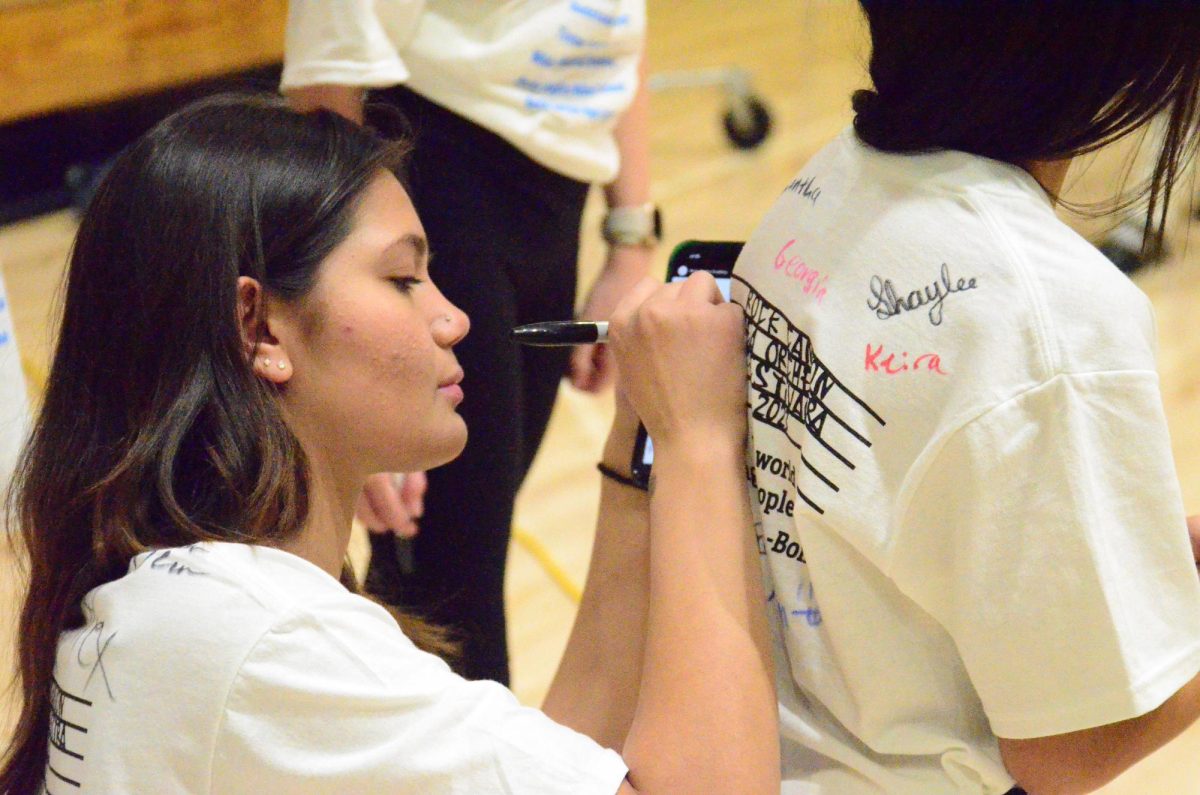 Sophia Lévesque ‘25 signs her name on the back of an elementary student’s shirt before the annual orchestra Feeder Festival Dec 7. The festival was a chance for elementary school and middle school students to see what being in high school orchestra is like, with over 200 musicians in the gym. “I love giving younger children the joy of seeing the huge high school and giving them high hopes for their futures in orchestra,” Lévesque said.
