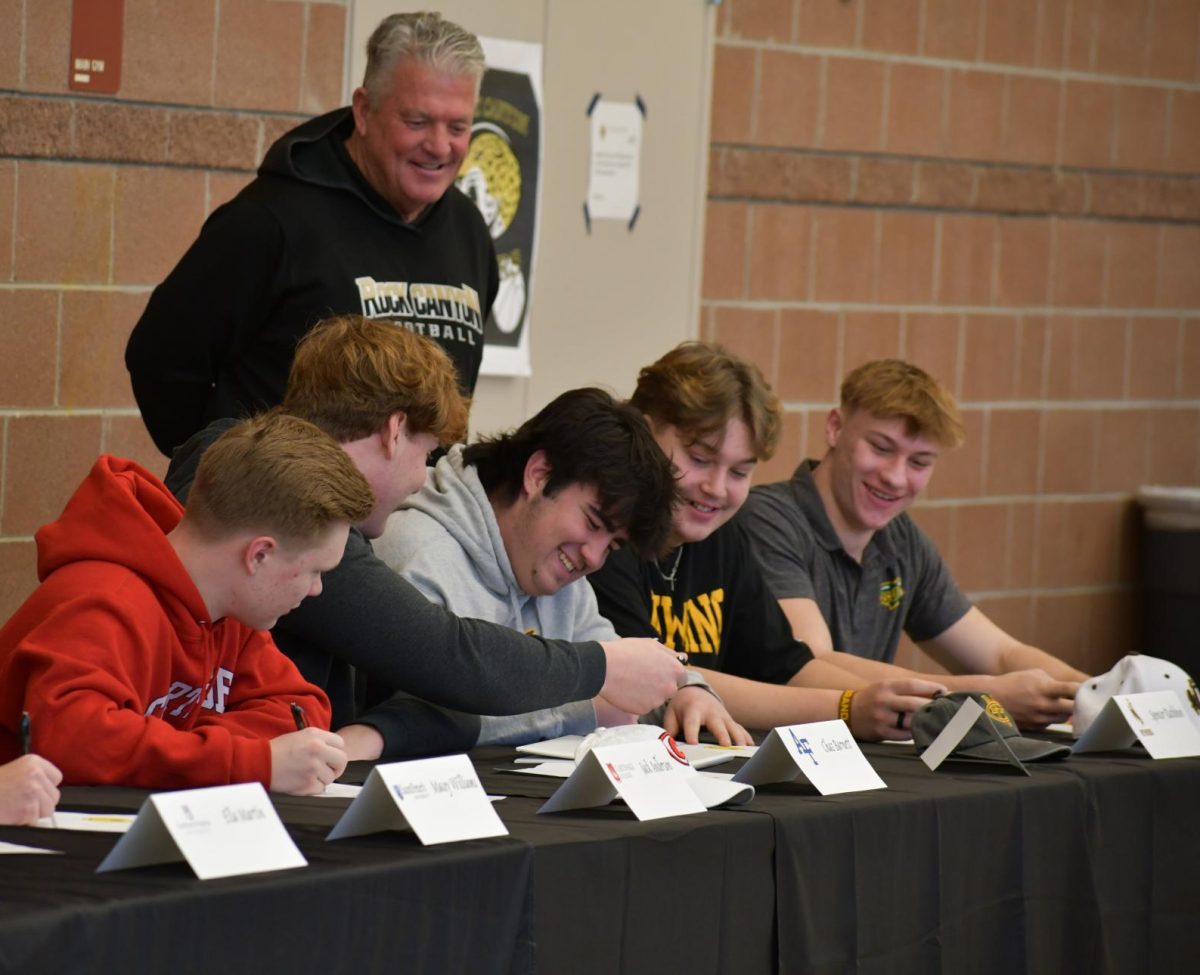 Head football coach Kevin Meyer stands over commits Jack Anderson ‘24, Chaz Barnett ‘24, Jack Munro ‘24, Spencer Rathbun ‘24 and Will Rosenmeyer ‘24 Feb. 7. Each football players all signed to a different school. Three other commits, along with Munro all signed to play for Colorado Mesa University (CMU): volleyball player Lindsay Heyliger ‘24, lacrosse player Aidan Brock ‘24 and basketball player Sienna Pillsbury ‘24 . 